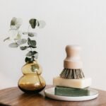 Composition of shaving brush placed on organic soaps on ceramic white holder near yellow transparent vase with plant on wooden round table against white wall