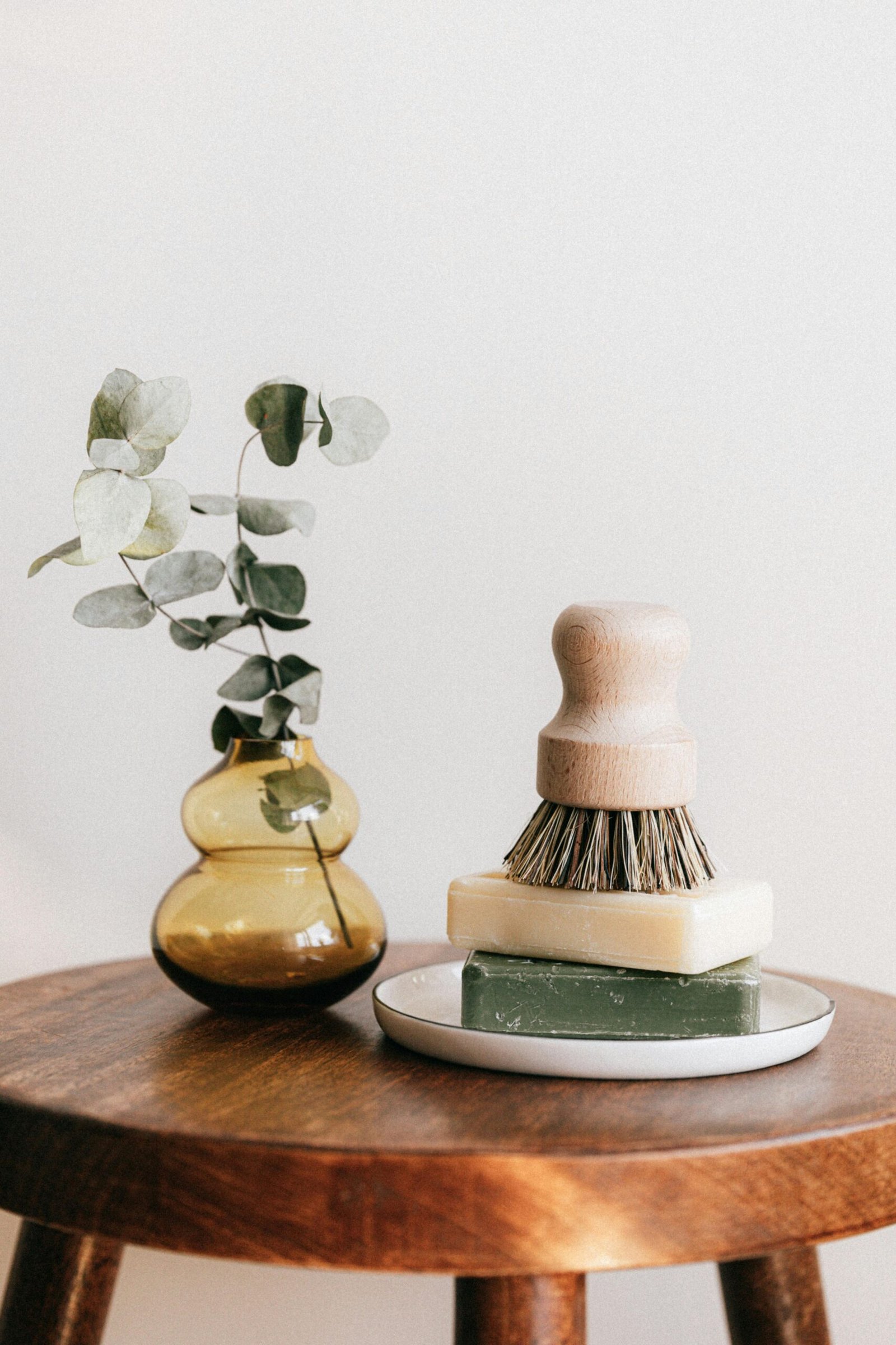 Composition of shaving brush placed on organic soaps on ceramic white holder near yellow transparent vase with plant on wooden round table against white wall